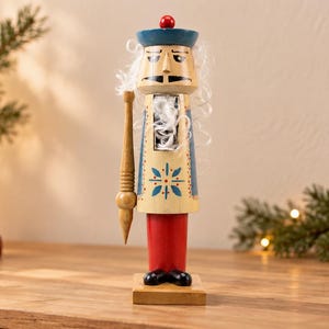 May include: A wooden nutcracker figurine featuring a blue hat with a red top knob, white hair, and a cream-colored torso decorated with a blue and red star pattern. The figure has red legs, black feet, a square base, and a tall wooden staff accessory.