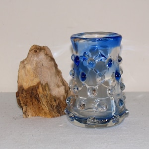 May include: A small, clear glass vase with a blue rim and blue and clear glass bubbles. The vase is sitting on a white surface.