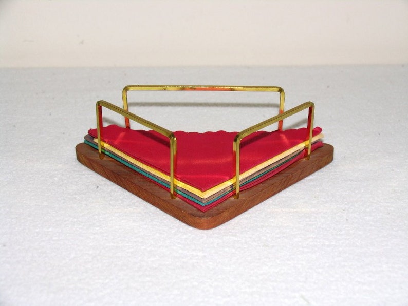 May include: A wooden napkin holder with a gold metal frame. The holder is triangular in shape and has a stack of colorful napkins inside.