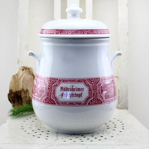 Puede incluir: Olla de cerámica blanca con tapa, detalles florales y texto en rojo. La olla tiene dos asas y una banda decorativa con las palabras "Rüdesheimer Früchtetopf". La tapa tiene un pequeño pomo. Un artículo de cocina vintage.