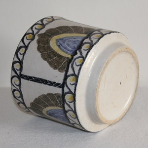 May include: A cylindrical ceramic pot with a white base and a decorative pattern. The pattern features stylized floral motifs in brown, blue, and yellow against a white background. The rim is adorned with a black and yellow border.