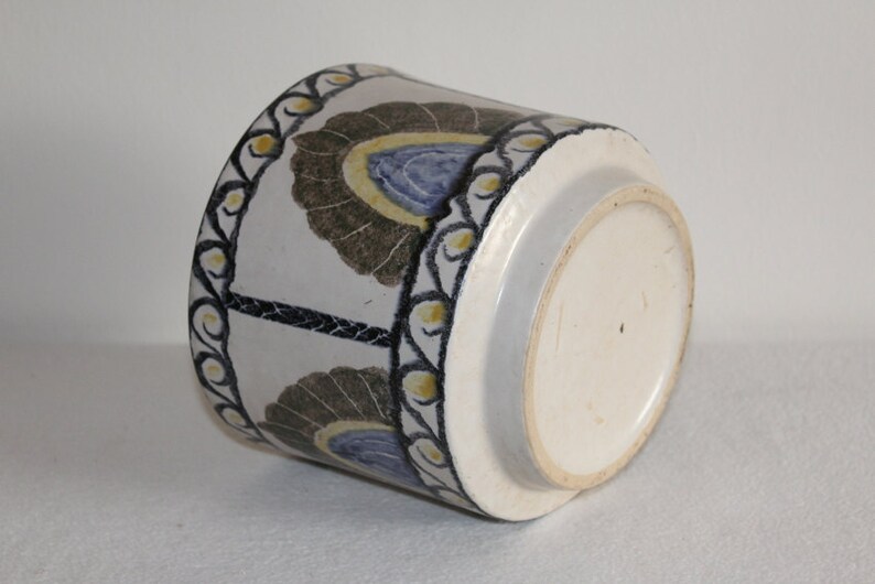 May include: A cylindrical ceramic pot with a white base and a decorative pattern. The pattern features stylized floral motifs in brown, blue, and yellow against a white background. The rim is adorned with a black and yellow border.