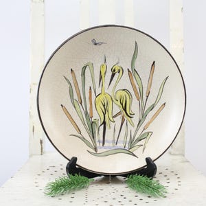 Könnte beinhalten: Dekorative Keramikplatte mit einer Naturszene mit gelben Vögeln, Schilf und einem Schmetterling. Die Platte hat einen cremefarbenen Hintergrund mit schwarzem Rand und wird auf einem schwarzen Ständer präsentiert.