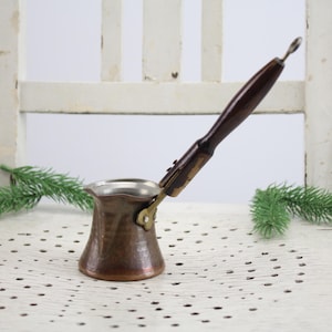 Könnte beinhalten: Eine kupferfarbene türkische Kaffeekanne, auch Cezve genannt, mit einem langen, dunklen Holzgriff. Die Kanne hat einen breiten Boden, der sich zu einem schmalen Hals verjüngt, und einen silberfarbenen Innenraum. Der Griff ist mit einem Messingscharnier befestigt.