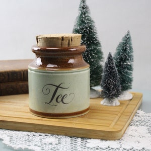 Puede incluir: Un tarro de té de cerámica marrón y verde con tapa de corcho. El tarro tiene la palabra "Tea" escrita en cursiva.