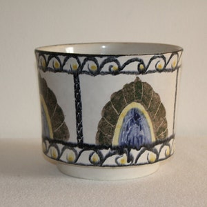 May include: A white ceramic planter with a decorative design. The planter features a repeating pattern of stylized fan shapes in brown, blue, and yellow. The rim and vertical lines are outlined in black.