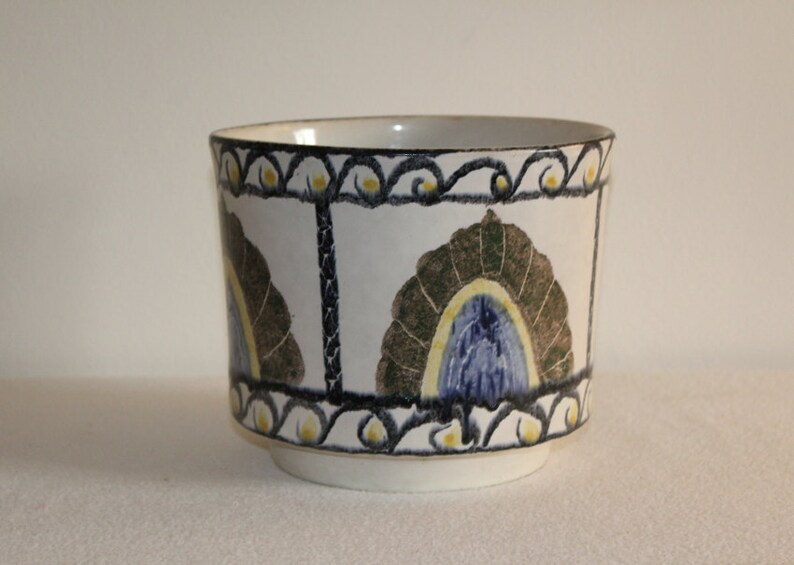 May include: A white ceramic planter with a decorative design. The planter features a repeating pattern of stylized fan shapes in brown, blue, and yellow. The rim and vertical lines are outlined in black.