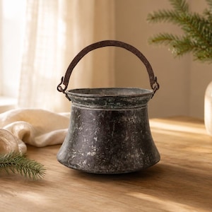 Puede incluir: Un cubo de metal oscuro con un asa curva, que muestra signos de desgaste. Se coloca sobre una superficie de madera, junto a un jarrón lleno de vegetación y una vela encendida. La escena evoca una sensación de calidez y es un objeto decorativo.