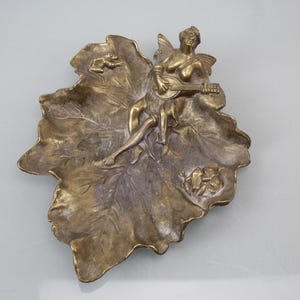 Puede incluir: Un plato decorativo de color bronce en forma de hoja grande. Una figura esculpida de una mujer con alas tocando un instrumento de cuerda está centrada en la hoja. El plato tiene una superficie texturizada detallada y una pequeña rana esculpida.