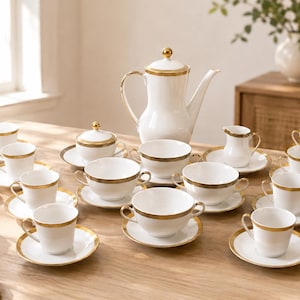 Puede incluir: Un juego de té de porcelana blanca que incluye una tetera alta con tapa de borde dorado, un azucarero con tapa, una jarra para leche y varias tazas con sus platos. Cada pieza presenta una fina banda dorada a lo largo del borde superior.