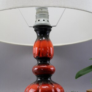 May include: A ceramic table lamp with a white lampshade. The lamp base is a glossy red and black colour. The base has a bulbous shape with a textured surface. The lamp is a decorative home decor item.