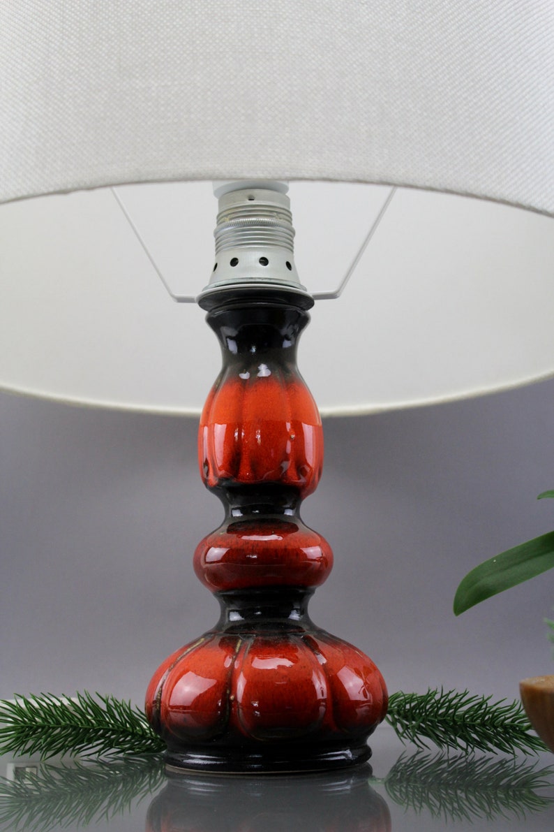 May include: A ceramic table lamp with a white lampshade. The lamp base is a glossy red and black colour. The base has a bulbous shape with a textured surface. The lamp is a decorative home decor item.