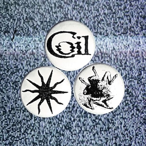 May include: Three white button pins with black designs. The first pin has the word "Gil" in a stylized font. The second pin has a black starburst design. The third pin has a black and white illustration of a creature with a long tail and legs.