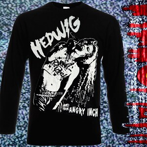 以下が含まれることがあります： 白いグラフィックプリントが施された黒い長袖Tシャツ。プリントには人物と「Hedwig and the Angry Inch」の文字が描かれている。