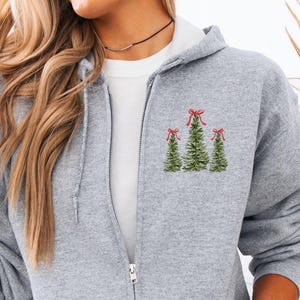 May include: A light gray zip-up hoodie with a white interior lining. The hoodie features an embroidered design of three Christmas trees with red bows on the left chest. The model is wearing light blue jeans.