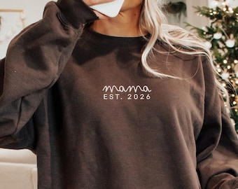 May include: A dark brown long-sleeve crewneck sweatshirt featuring the white embroidered text "mama" in a cursive script above the printed text "EST. 2026" in a clean, sans-serif font centered on the upper chest area of the garment.