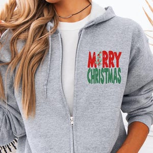 May include: A light gray zip-up hoodie with the words "MERRY CHRISTMAS" in red and green. The hoodie has a white interior and a front pocket. The model is wearing light blue jeans.