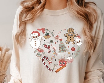 May include: A cream-colored long-sleeved sweatshirt with a heart-shaped design featuring various Christmas-themed illustrations, including a snowman, a gingerbread man, a Christmas tree, candy canes, and a mug of hot chocolate.