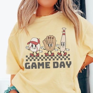 May include: A yellow t-shirt featuring a graphic of a baseball, glove, and bat with faces. The graphic is in a retro style and says "GAME DAY" underneath.