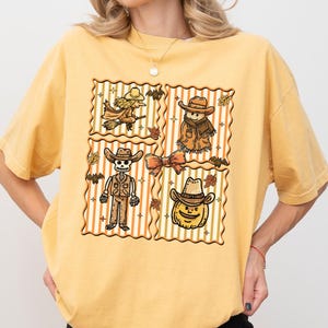 May include: Mustard yellow t-shirt with a four-panel graphic of Halloween cowboy illustrations. Each panel features a different character: a witch, a scarecrow, a skeleton, and a pumpkin, all wearing cowboy hats.