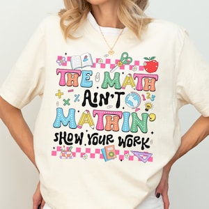 May include: Cream-colored t-shirt with the text "THE MATH AIN'T MATHIN' SHOW YOUR WORK" in colorful, playful lettering. The design includes math symbols, an apple, scissors, and other school-related graphics. The shirt is paired with black shorts.