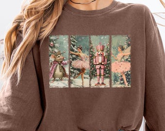 May include: A brown sweatshirt featuring a rectangular graphic of The Nutcracker ballet. The graphic shows a mouse, a ballerina, a nutcracker, and another ballerina in a winter scene. The sweatshirt is worn by a person wearing denim shorts.