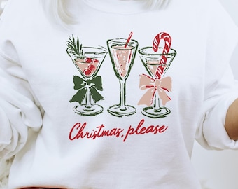 May include: A white long-sleeve sweatshirt featuring a graphic print of three cocktail glasses decorated with a green bow, a pink bow, and a candy cane. The text "Christmas, please" is printed in red script below the glasses.