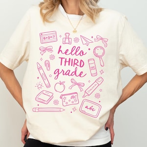 May include: Cream-colored t-shirt with a pink graphic design featuring school-related illustrations and the text "hello THIRD grade". The design includes a crayon, apple, pencil, and other school supplies. The shirt is a casual, comfortable style.