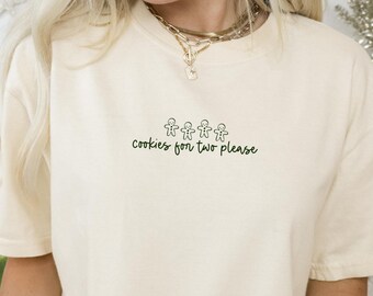 May include: A cream-colored short-sleeve t-shirt featuring a small graphic of four gingerbread men in a row above the dark green text that reads, "cookies for two please." The shirt has a crew neckline and a relaxed, casual fit.