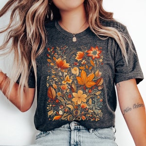 May include: A dark gray t-shirt with a floral print in shades of orange, yellow, and green. The flowers are arranged in a bouquet-like shape and are surrounded by leaves and vines.
