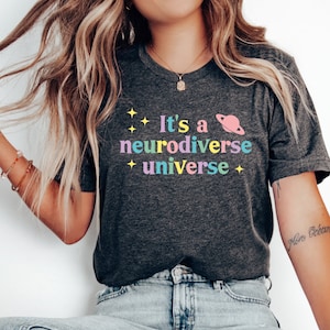Pu&ograve; includere: Una maglietta grigio scuro con il testo "It's a neurodiverse universe" in colori arcobaleno. Il testo &egrave; circondato da piccole stelle e un pianeta rosa con un anello.