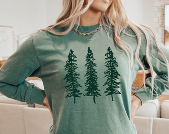 May include: A long-sleeved, green shirt featuring a dark green graphic of three stylized pine trees. The shirt is paired with gray shorts. The overall aesthetic suggests a casual, nature-inspired theme.