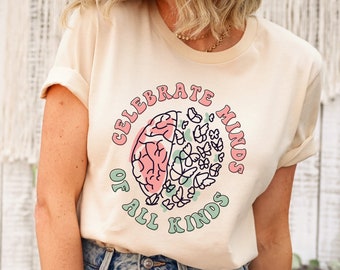 May include: A light beige t-shirt with a graphic of a brain with flowers and the text "Celebrate Minds of All Kinds".