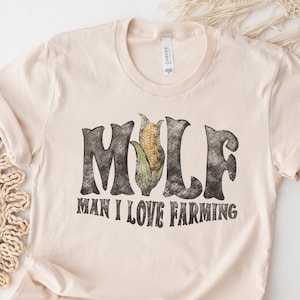 May include: A beige t-shirt with a distressed black graphic that says "MULF Man I Love Farming" with an ear of corn in the middle of the word "MULF".