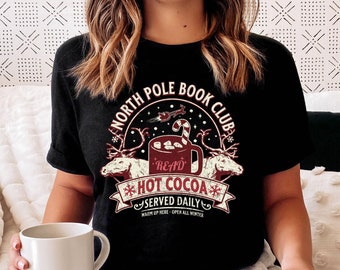 May include: A black t-shirt with a white graphic design that says "North Pole Book Club" and features a mug of hot cocoa with marshmallows and a candy cane. The text "Read Hot Cocoa" is written in red on the mug. The graphic also includes a reindeer and snowflakes. The text "Served Daily" is written below the mug.