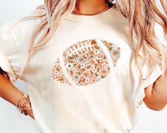 May include: Ivory-colored t-shirt featuring a floral football design in shades of brown, peach, and white. The shirt has short sleeves and is paired with blue jeans. The overall style is casual and relaxed.