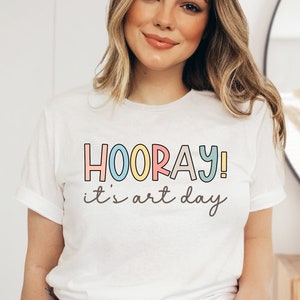 May include: White t-shirt with the text "HOORAY! it's art day" in colorful letters.