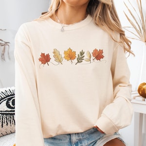 May include: Cream-colored long-sleeve shirt featuring a line of embroidered autumn leaves in shades of red, yellow, and green. The shirt is made of a soft material and is suitable for fall. The model is wearing denim shorts.