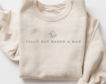 May include: A cream-colored crewneck sweatshirt featuring a small embroidered Santa hat above the text JOLLY BUT NEEDS A NAP. The word JOLLY is stitched in red, while the remaining words are in black, centered on the chest of the garment.