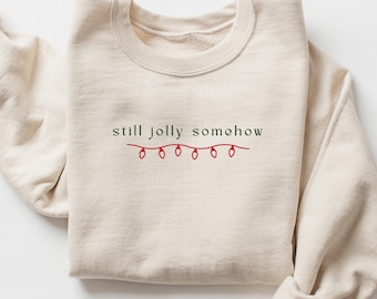 May include: A cream-colored crewneck sweatshirt featuring the dark green text "still jolly somehow" embroidered on the chest, positioned above a simple red line drawing of a string of five small Christmas light bulbs.