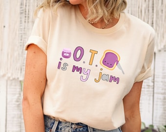 May include: A light beige t-shirt with a graphic of a jar of jam and a slice of bread with a smiling face. The text on the shirt reads "O.T. is my jam".