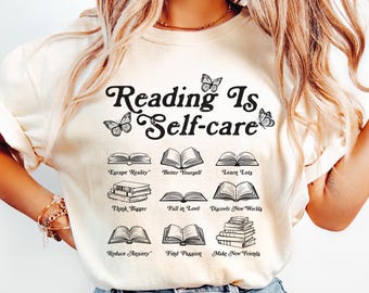 May include: A white t-shirt with the text "Reading Is Self-care" in a black script font. The shirt features a series of black and white illustrations of books with text below each book, such as "Escape Reality", "Better Yourself", "Learn Lots", "Think Bigger", "Fall in Love", "Discover New Worlds", "Reduce Anxiety", "Find Passion", and "Make New Friends".