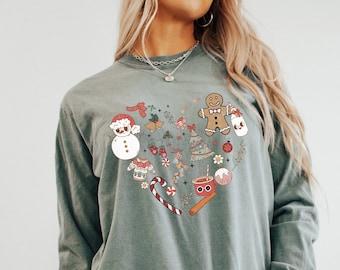 May include: A teal long-sleeve t-shirt with a heart-shaped design featuring various Christmas-themed illustrations, including a snowman, gingerbread man, candy canes, ornaments, and a Christmas tree.