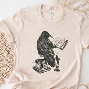 Puede incluir: Una camiseta beige con una ilustraci&oacute;n en blanco y negro de un cuervo posado sobre un libro, sosteniendo una pluma en su pico. El cuervo est&aacute; leyendo un libro con el texto "The Raven" visible.