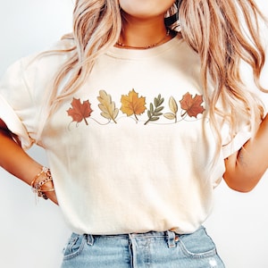 May include: A white t-shirt with a line of autumn leaves printed on the front. The leaves are in shades of red, orange, yellow, and green.