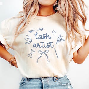 May include: A white t-shirt with the text "lash artist" in blue lettering. The shirt also features a blue illustration of eyelashes, a heart, a bow, and other lash-related tools.
