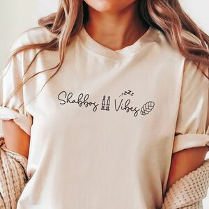 May include: A white t-shirt with the text "Shabbos Zzz Vibes" and a small illustration of a challah bread.