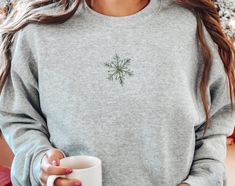 May include: A light gray crewneck sweatshirt featuring a small, embroidered dark green snowflake design centered on the chest. The garment has long sleeves and a ribbed neckline, providing a simple and clean look for the winter season.