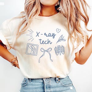 K&ouml;nnte beinhalten: Ein wei&szlig;es T-Shirt mit dem Text "X-ray tech" in blauer Schrift. Das Shirt zeigt au&szlig;erdem ein Herz, einen Sonnenstrahl, ein Blatt, eine R&ouml;ntgenaufnahme des Knies, eine Schleife und eine R&ouml;ntgenaufnahme des Brustkorbs.