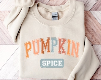May include: A beige sweatshirt with the text "PUMPKIN SPICE" in a rainbow arch design. The text is in a vintage collegiate font style.